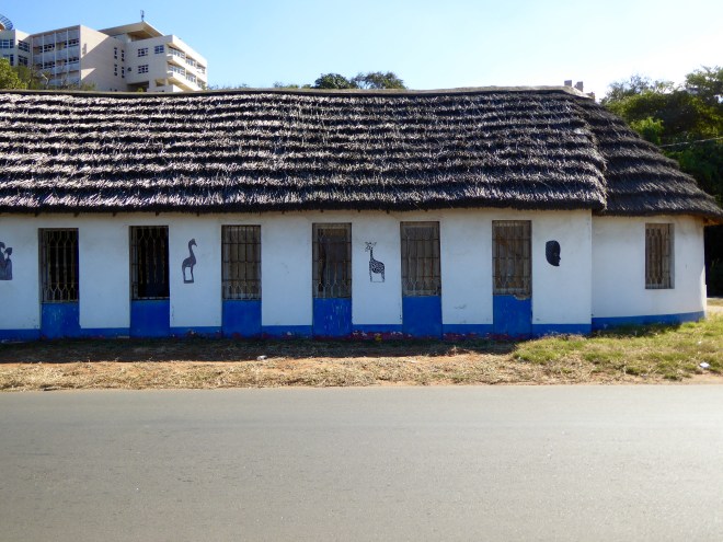 Old building with thatched roof. Zeb on ground under giraffe